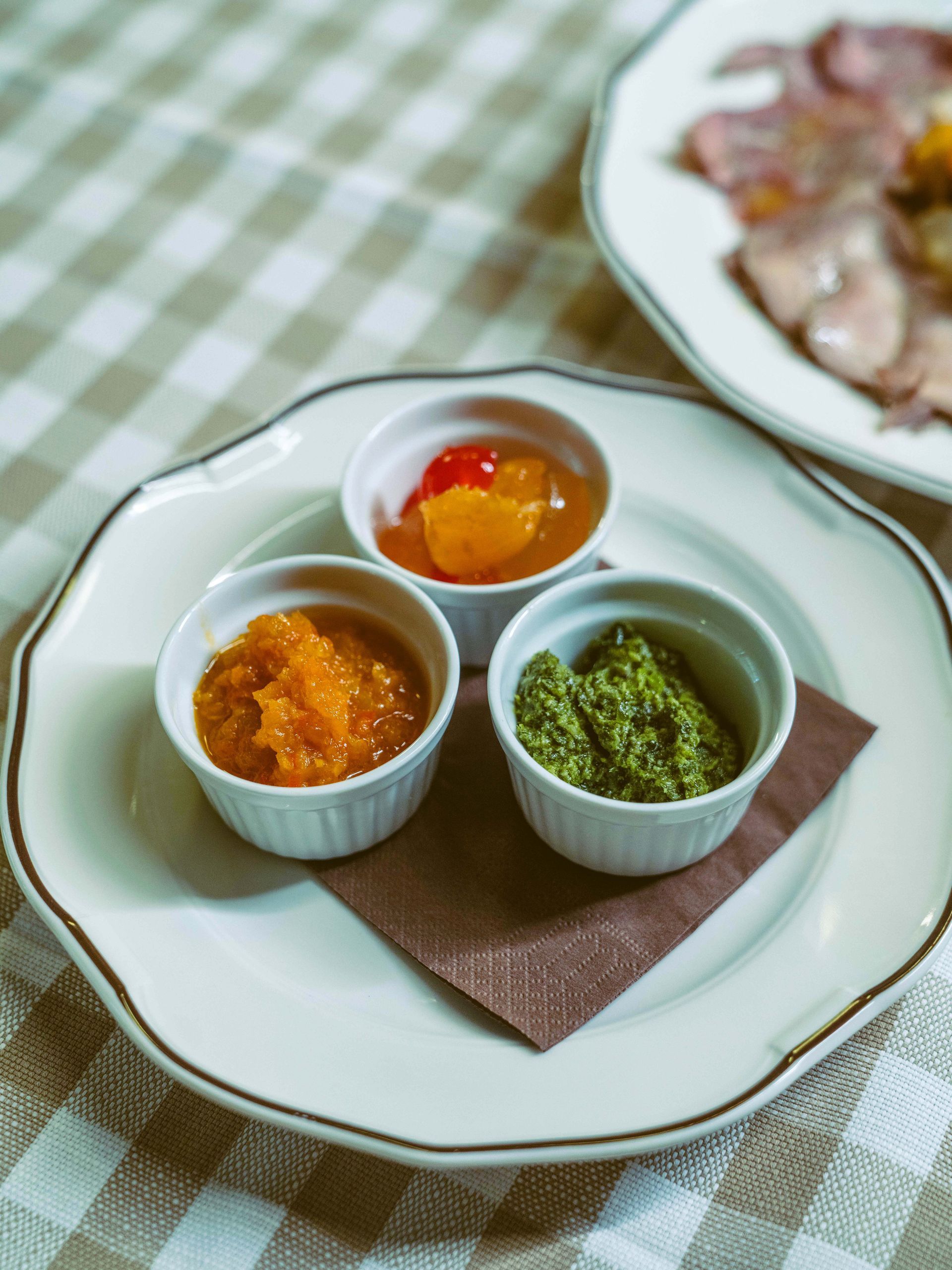 Piccoli piatti bianchi con salse arancioni, gialle e verdi su un piatto, disposti su una tovaglia a quadri.