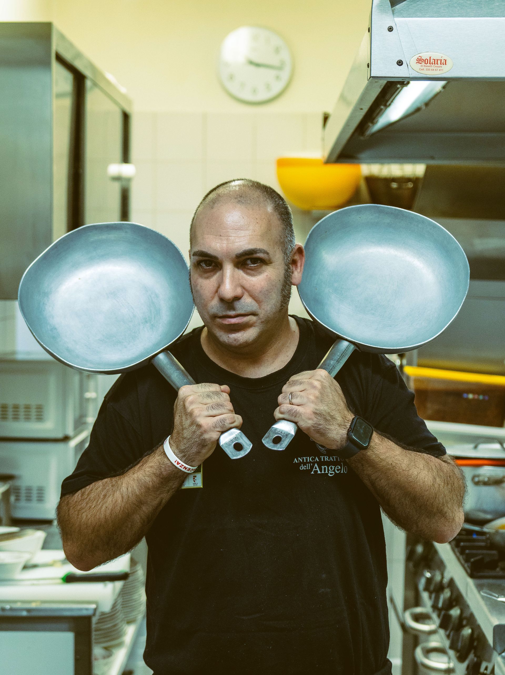 Cuoco in cucina con due padelle in mano e sguardo rivolto alla telecamera.