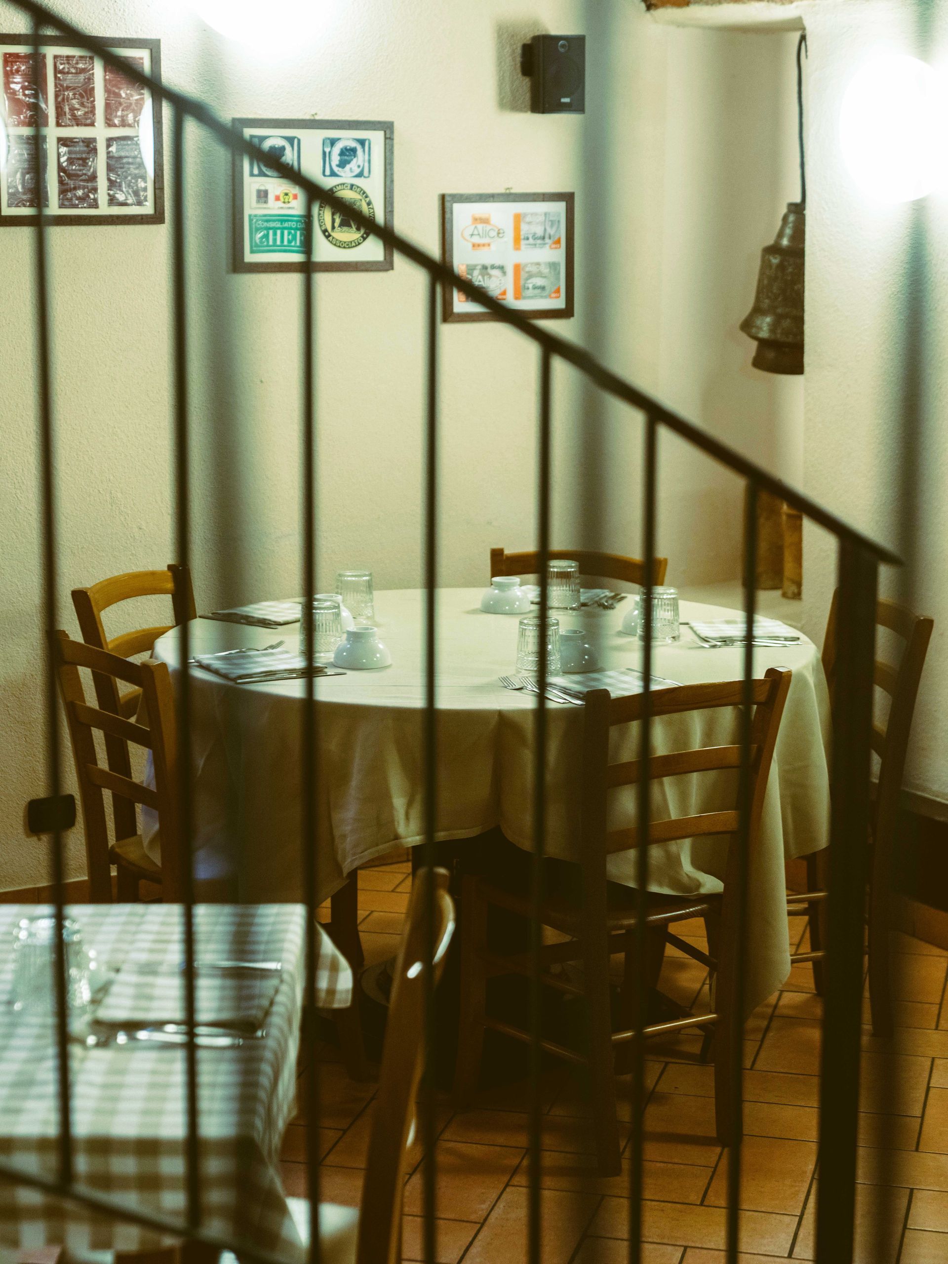 Sala da pranzo del ristorante con tavolo rotondo, sedie e opere d'arte incorniciate; vista attraverso la ringhiera di una scala.