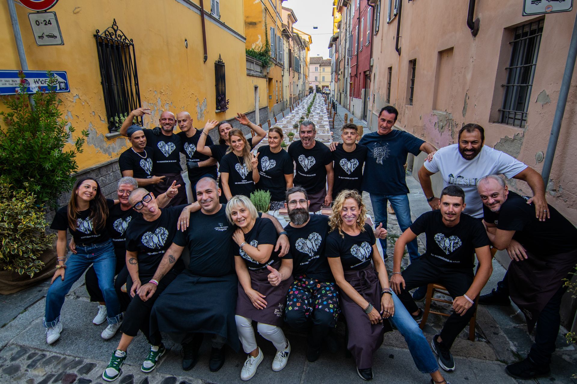 Un gruppo di persone, che indossano magliette abbinate con un disegno a forma di cuore, posa su una strada stretta e in pendenza con edifici colorati.