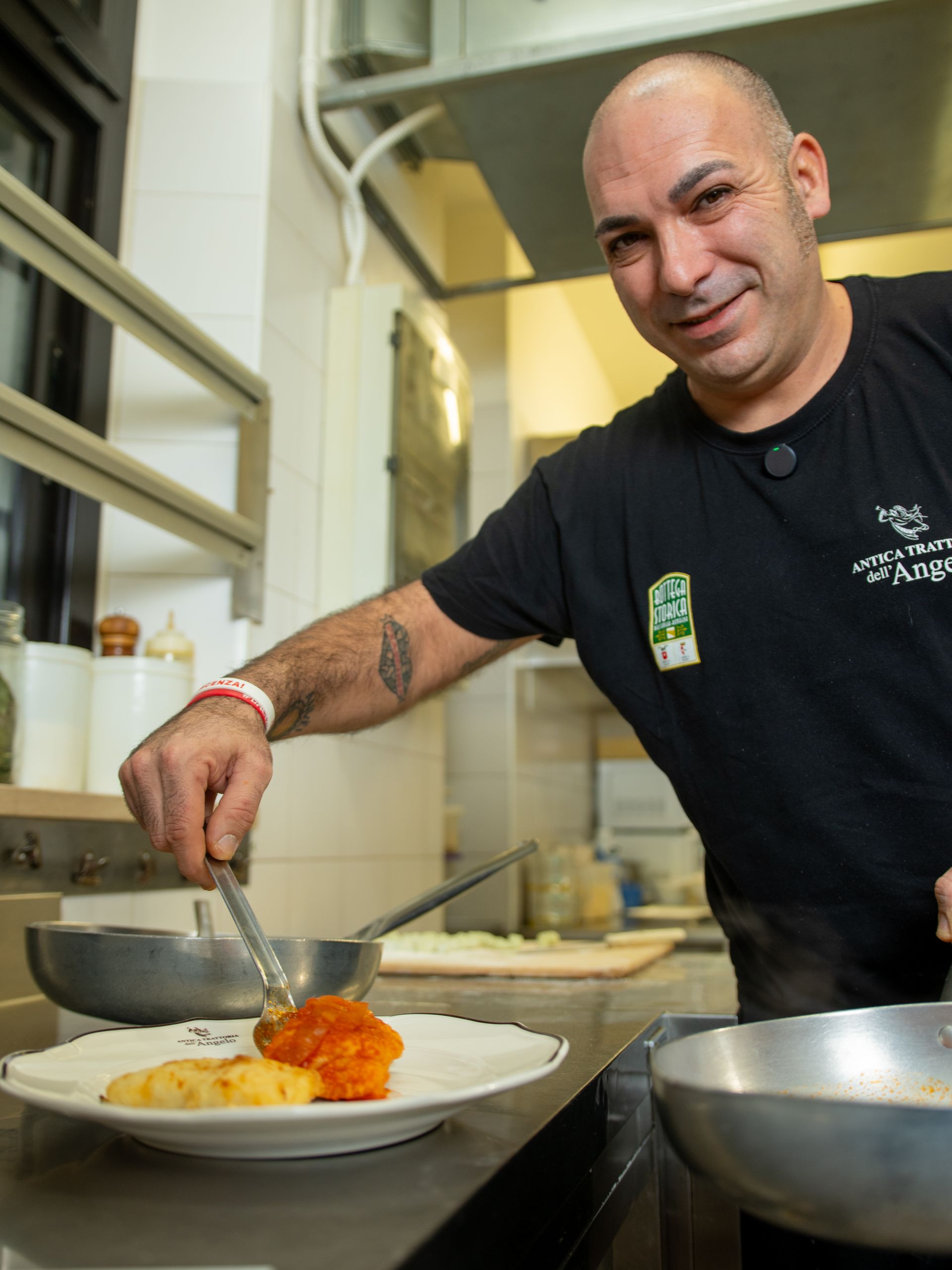 Uno chef impiatta il cibo in cucina. Sorride, indossa una camicia nera e ha un tatuaggio. Si vede un piatto con cibo arancione e giallo.