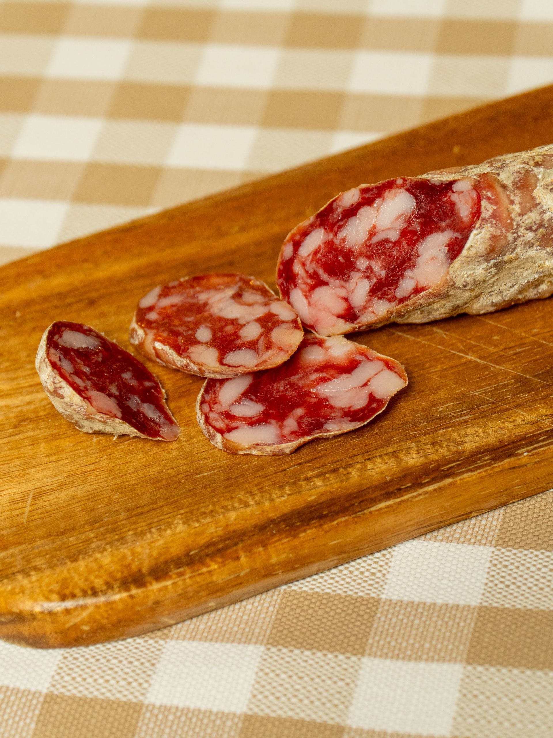 Fette di salame su un tagliere di legno, adagiate su una tovaglia a quadri.