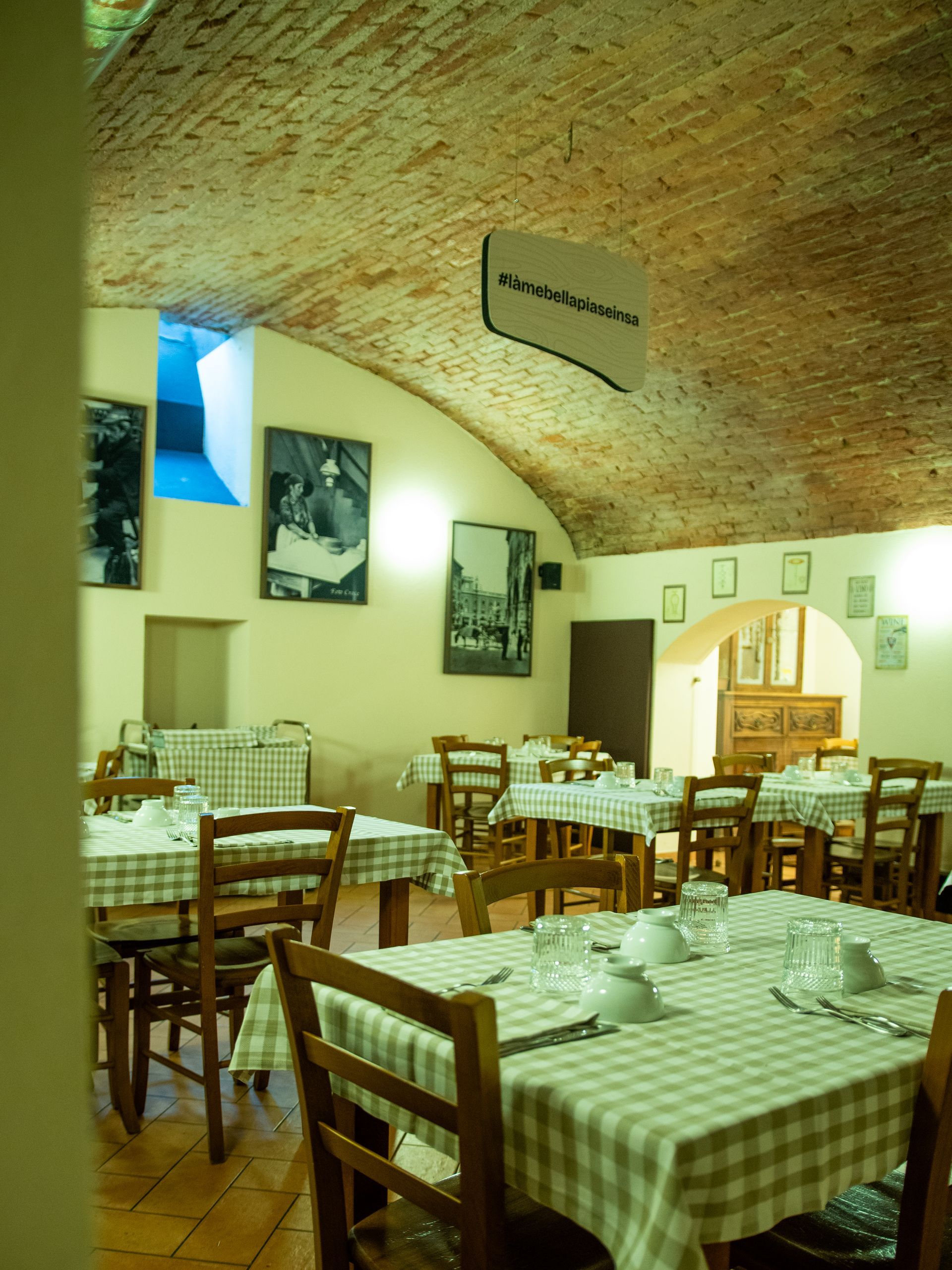 Interno del ristorante con tovaglie a quadri, sedie in legno, soffitto in mattoni e opere d'arte incorniciate alle pareti.