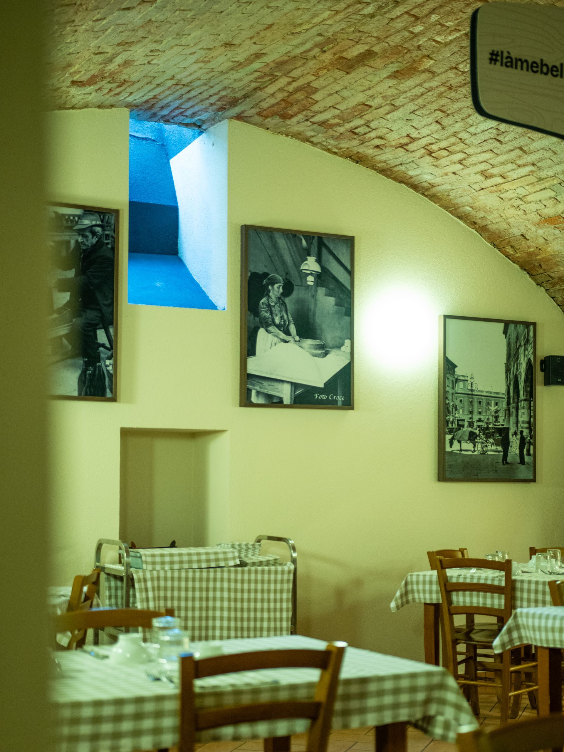 Interno del ristorante con soffitto a volta in mattoni, tavoli con tovaglie a quadri e foto in bianco e nero incorniciate alle pareti.