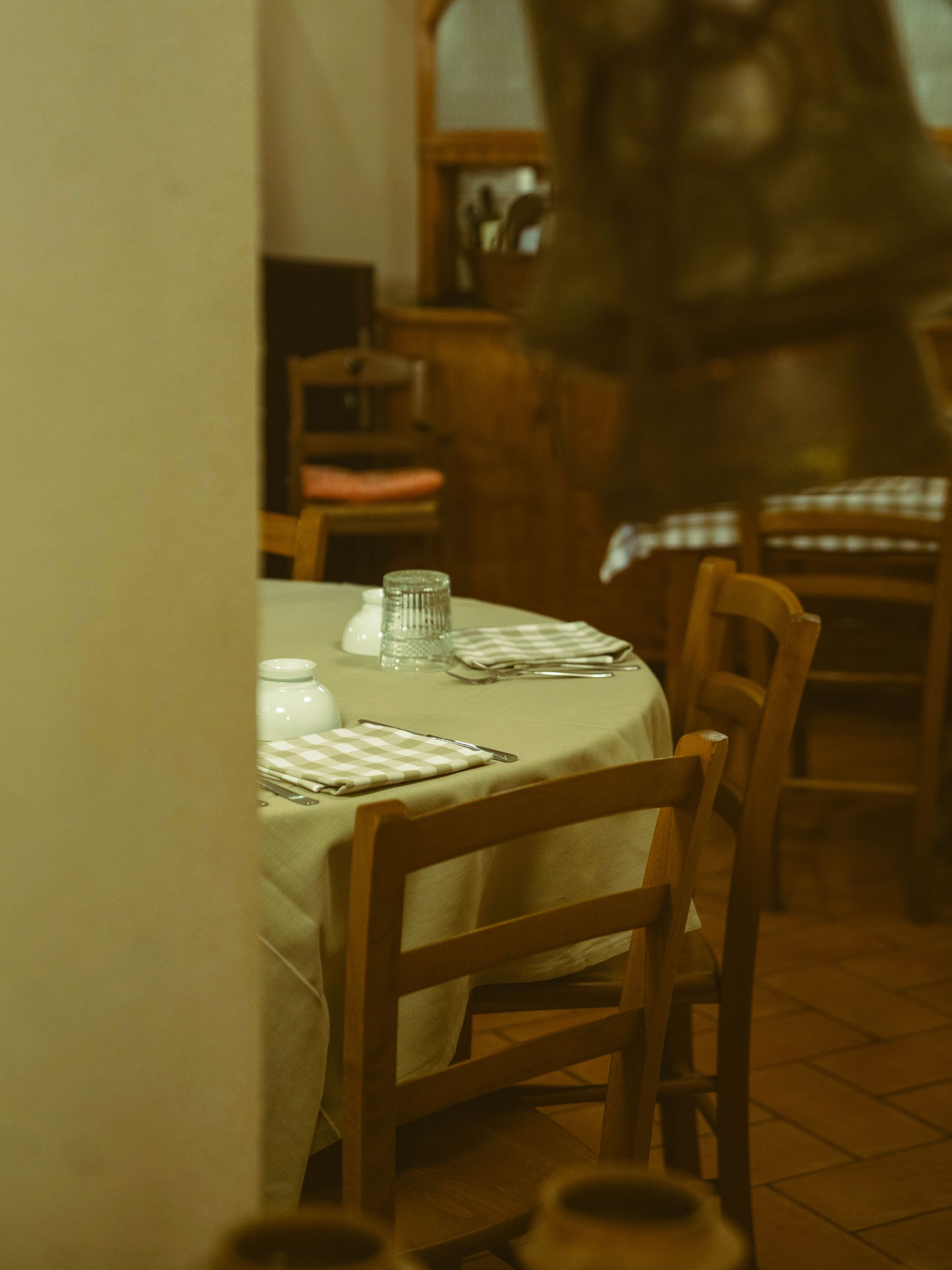 Tavolo rotondo apparecchiato per un pasto in un ristorante; due sedie di legno; piatti e bicchieri visibili.