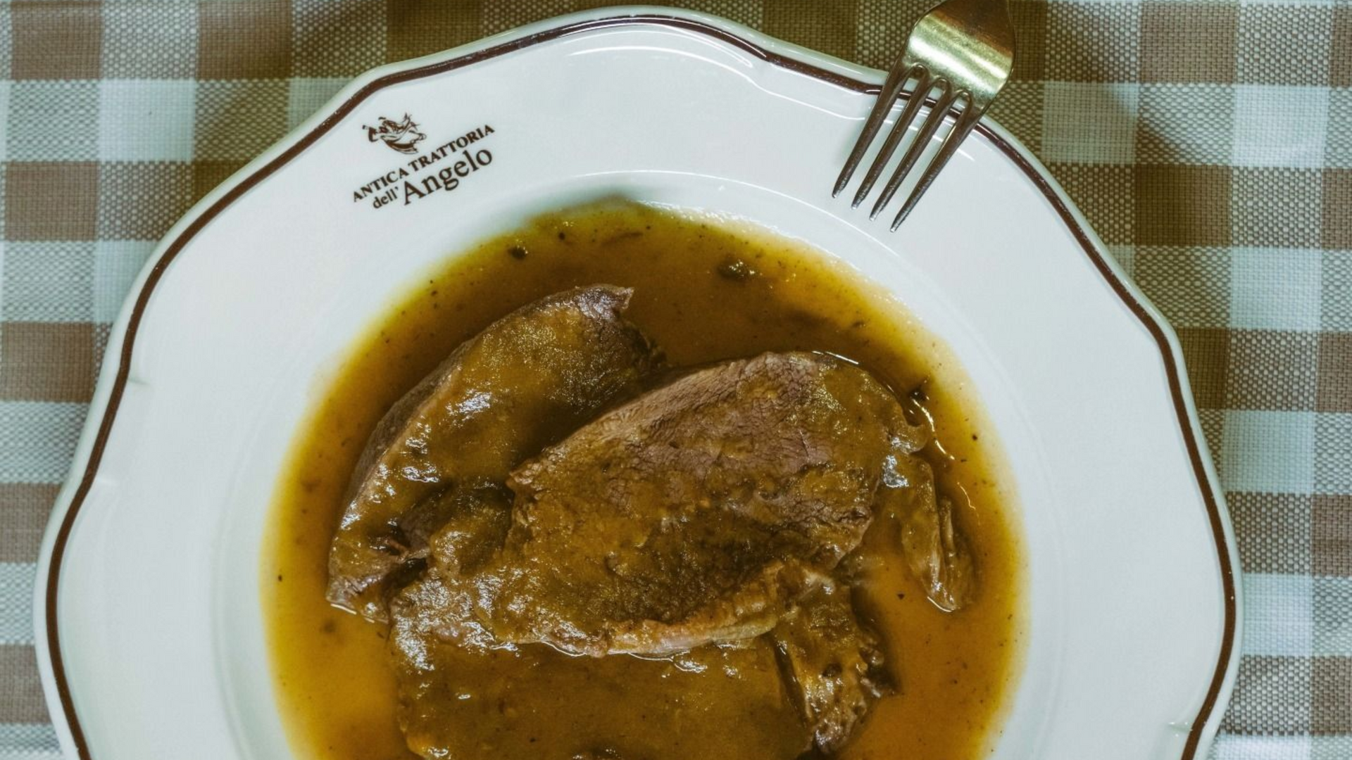 Tavolo apparecchiato per un pasto al ristorante: piatto di barbabietole, cestino del pane, bicchiere di vino e stoviglie su una tovaglia a quadri.