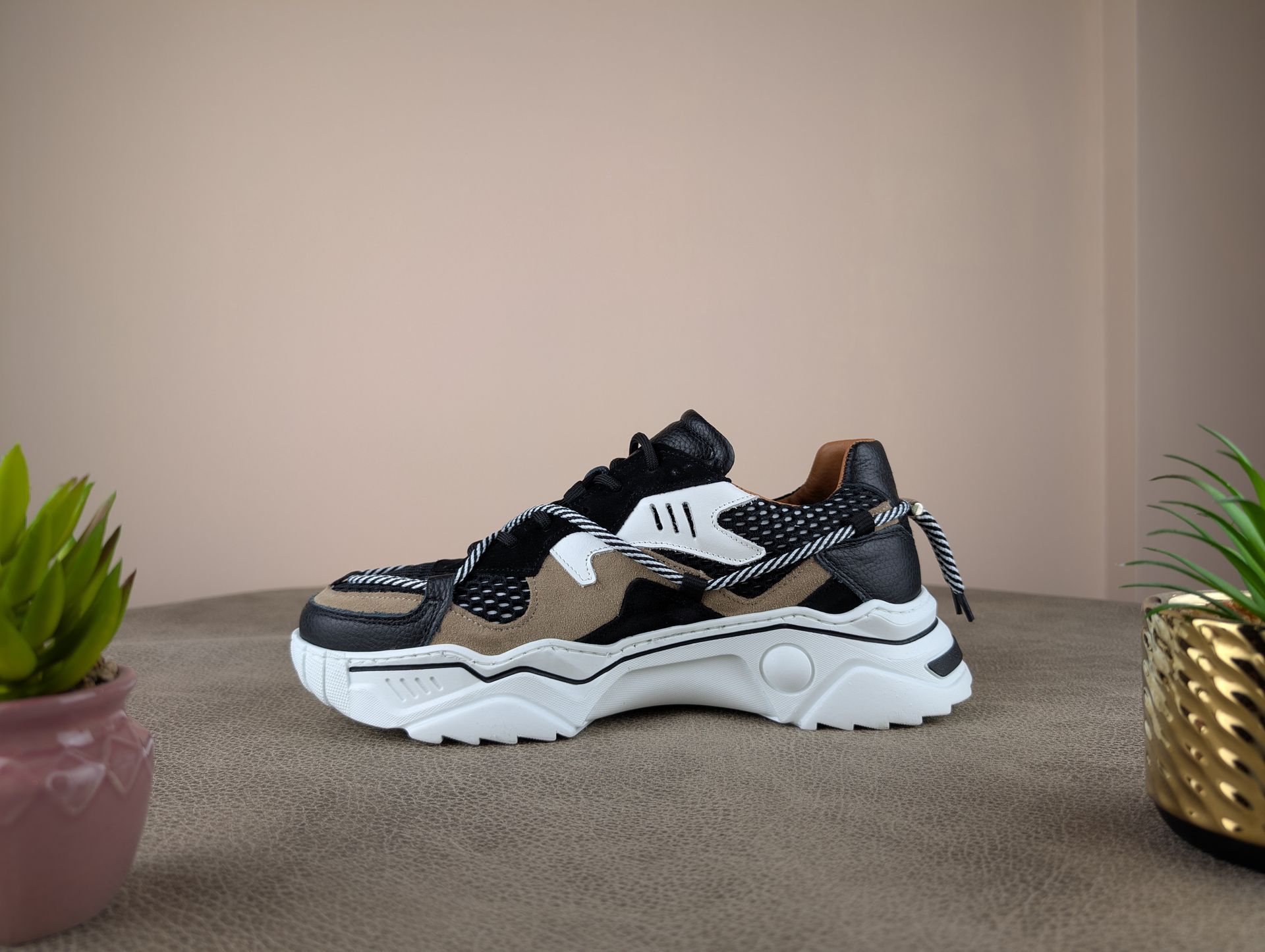 Stevige zwarte, bruine en witte sneaker op een gestructureerd oppervlak met kleine potplanten op de achtergrond.