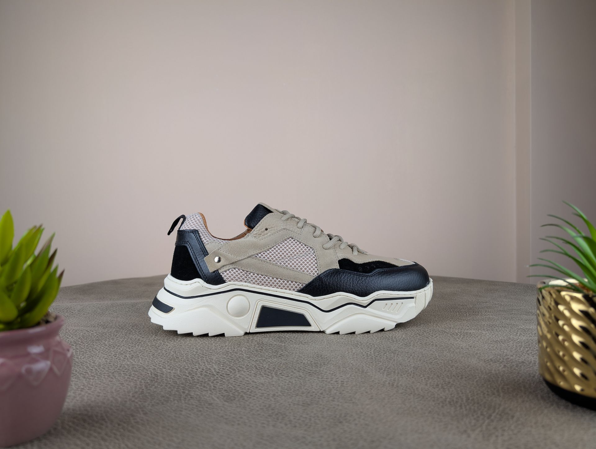 Stevige bruine, zwarte en witte sneakers op een gestructureerd oppervlak, geflankeerd door potplanten tegen een neutrale muur.