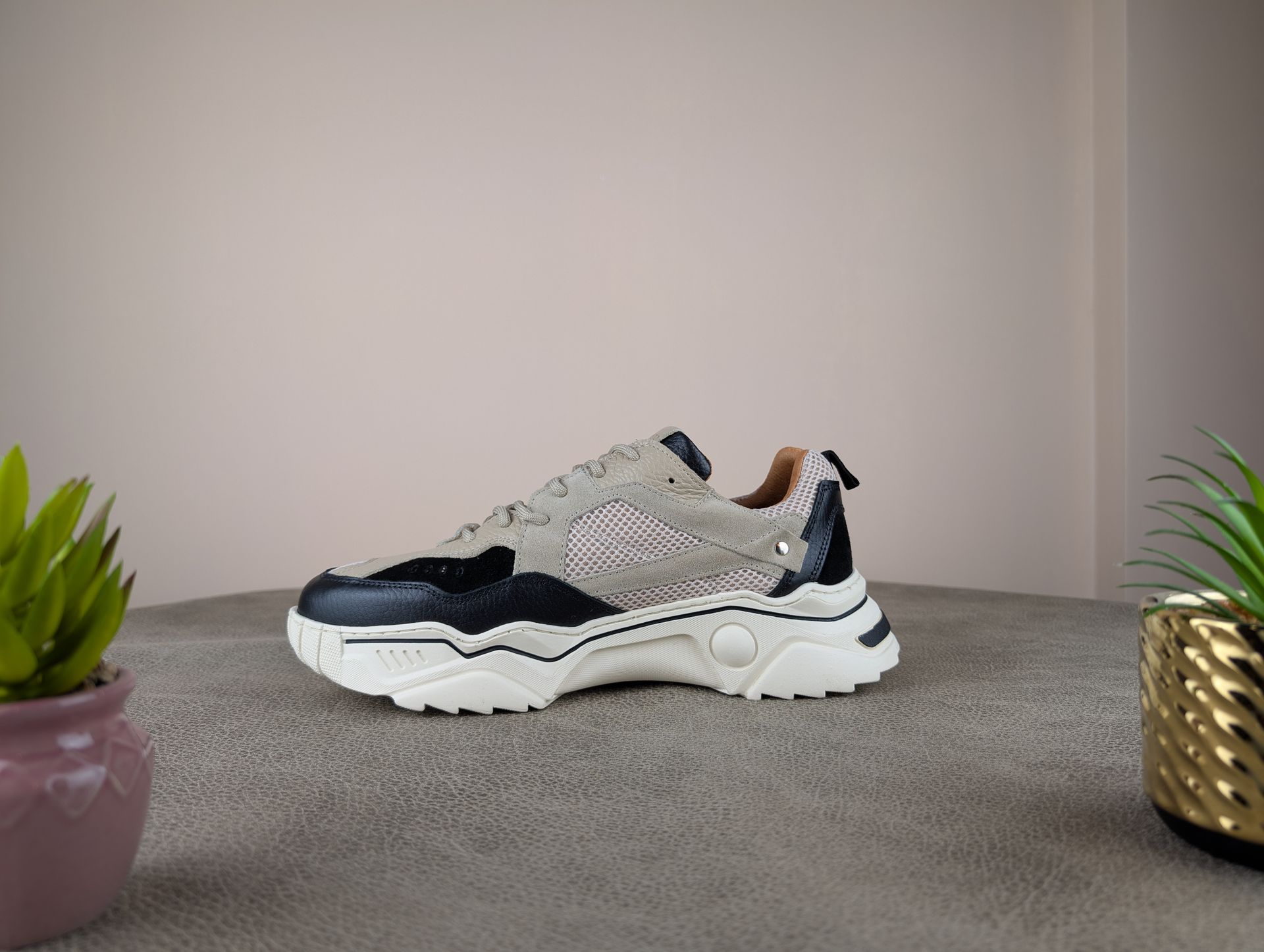 Stevige bruine, zwarte en witte sneakers op een gestructureerd oppervlak, geflankeerd door potplanten tegen een neutrale muur.