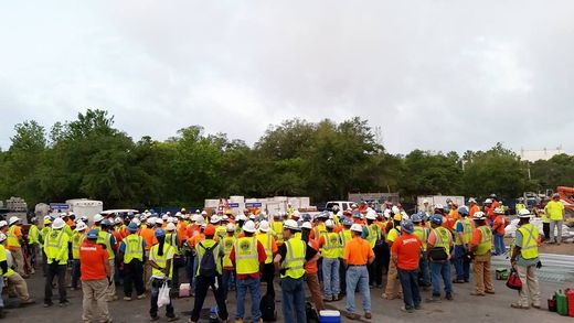 A large group of construction workers are standing in a parking lot.
