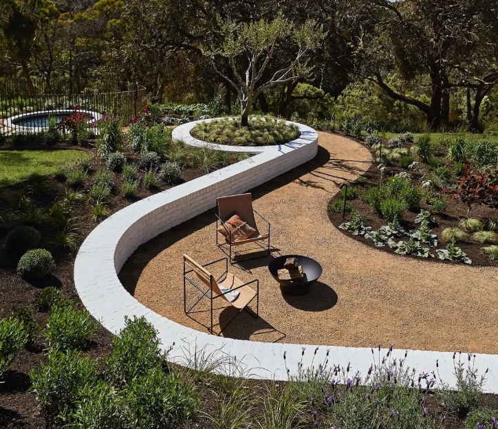 A curved walkway in a garden with two chairs and a fire pit