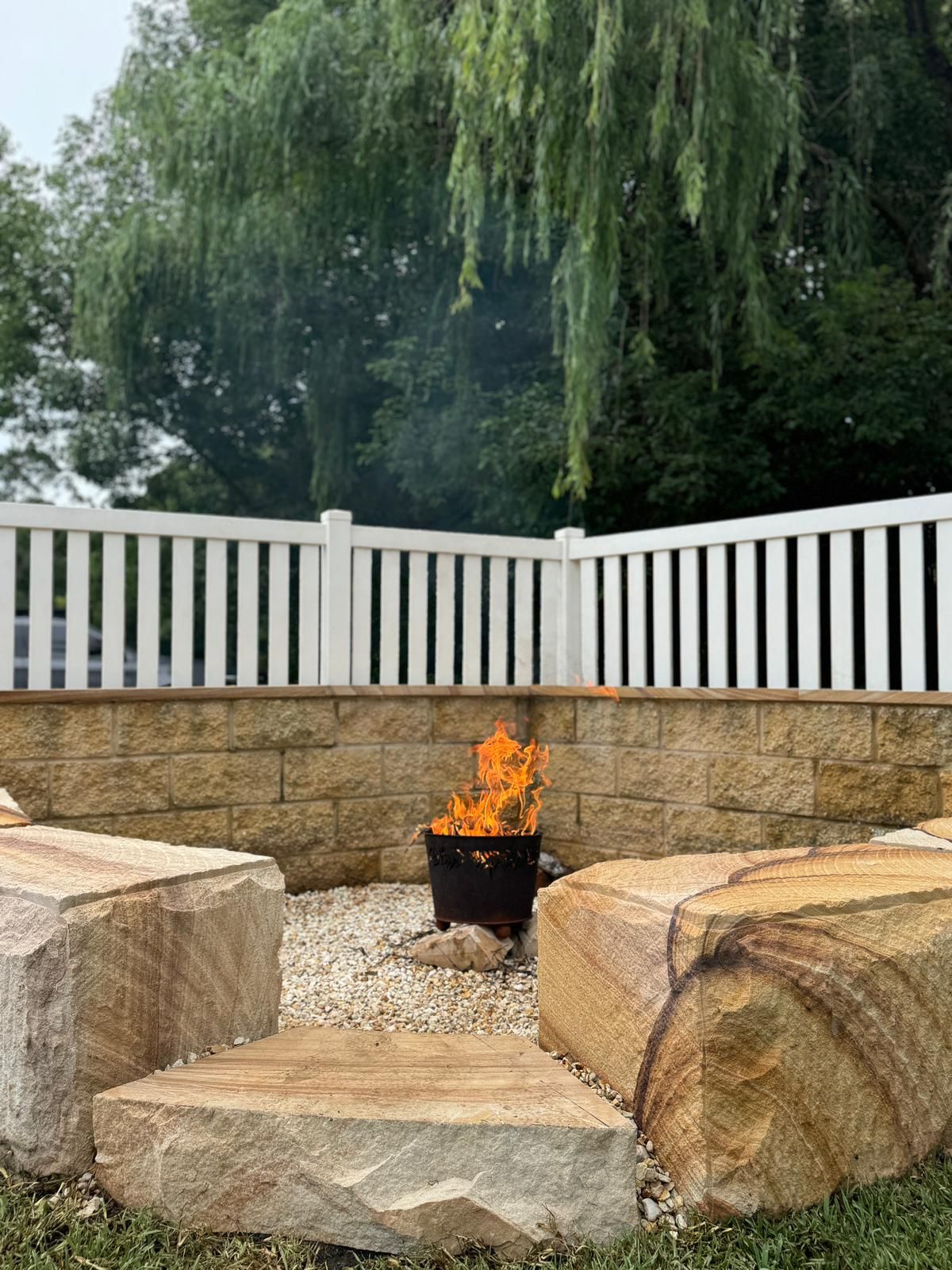 A fire pit is surrounded by rocks and a white fence.