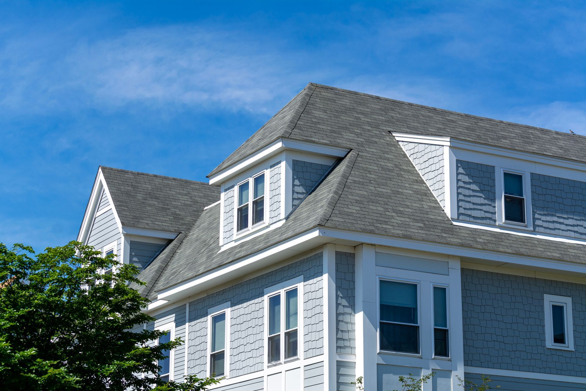 Blue-gray house with dormer windows, gray roof, and white trim against a blue sky, partially obscured by green tree.