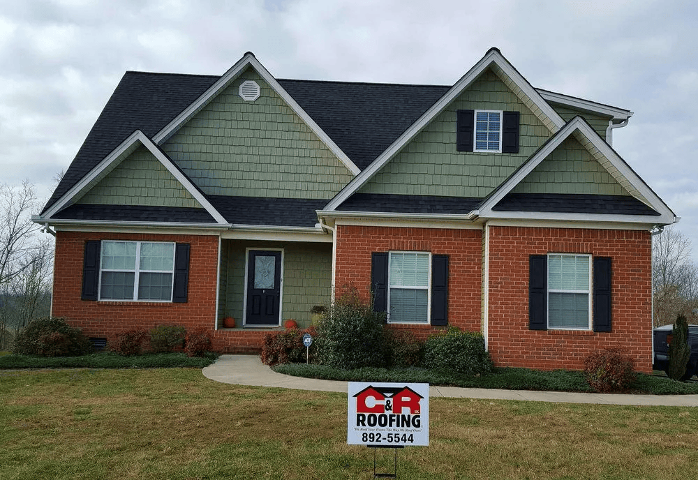 House with green siding and brick facade, black roof, and CR Roofing sign in yard.