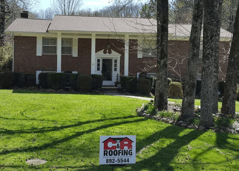 Red brick house with new roof, lawn, and a C&R Roofing sign.