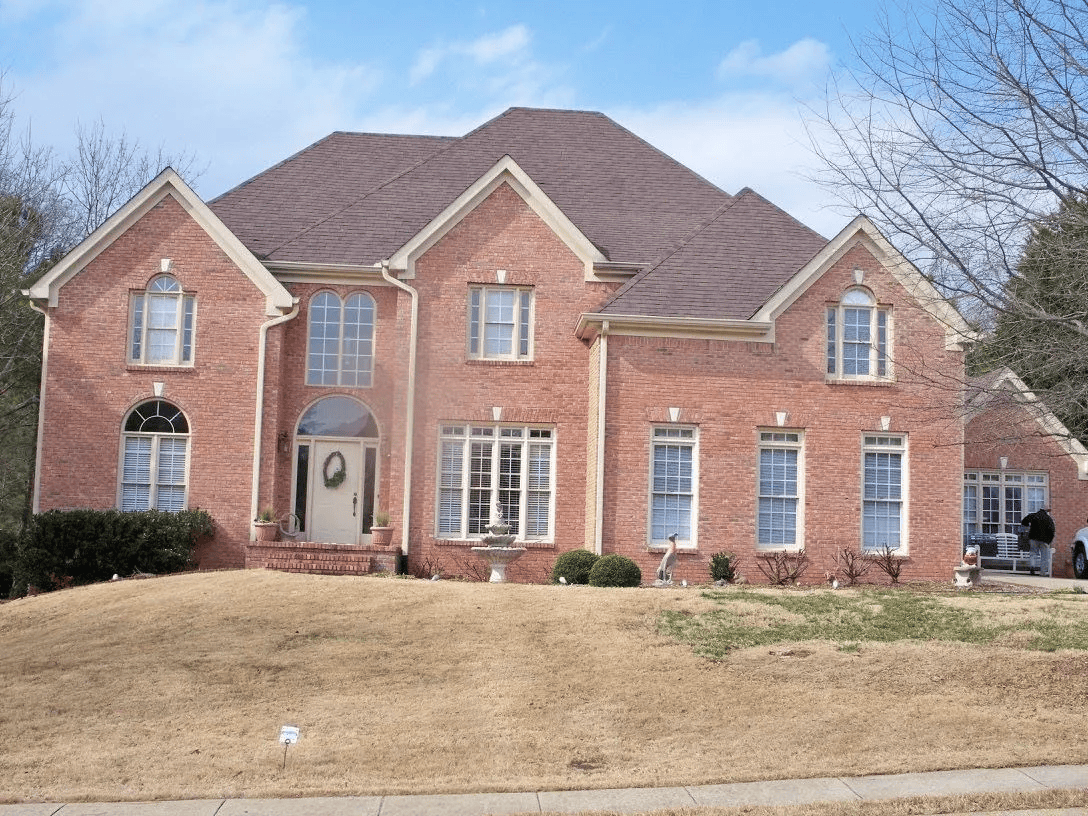 Red brick two-story house with a brown roof on a slight hill, with a blue sky background.