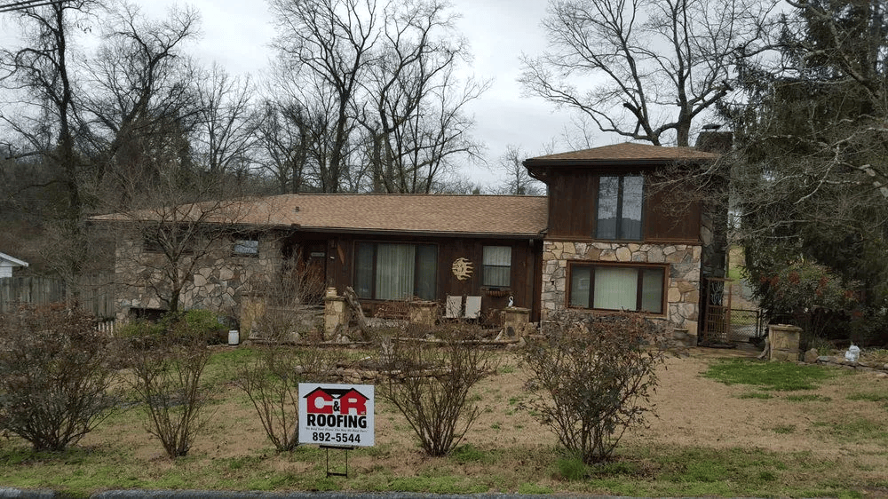 Stone-clad house with brown roof and chimney, surrounded by leafless trees and bushes; a sign is in the yard.