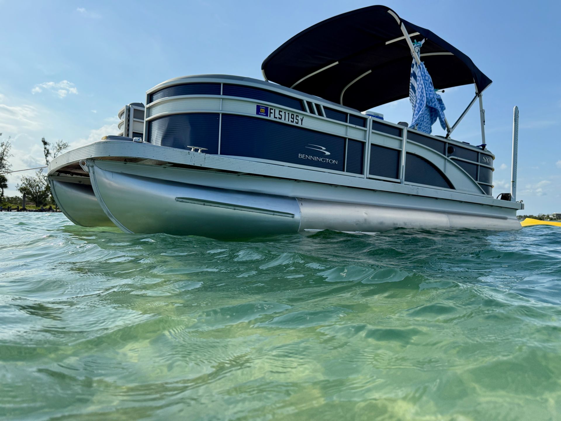 Pontoon boat on the water under a blue sky, with a dark canopy and flag.