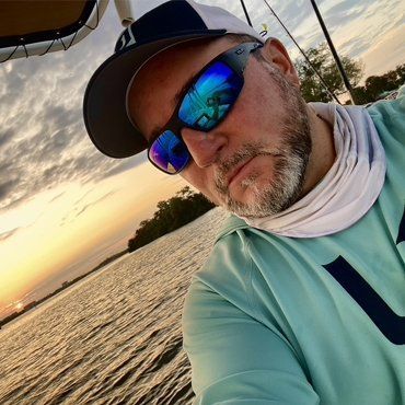 Man in sunglasses, hat, and neck gaiter on a boat at sunset.