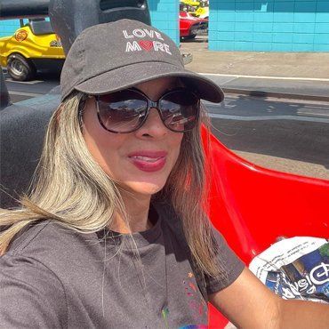 Woman in sunglasses and cap smiles, sitting in a red vehicle, possibly at an amusement park.