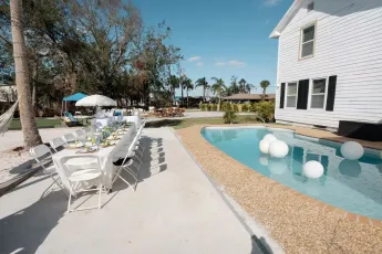 Backyard party setup with pool, long table, chairs, and white house. White balloons float in pool.