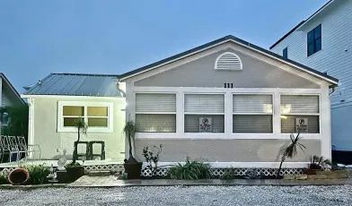 Small, light gray bungalow with white trim, three windows, and a smaller building to the left.