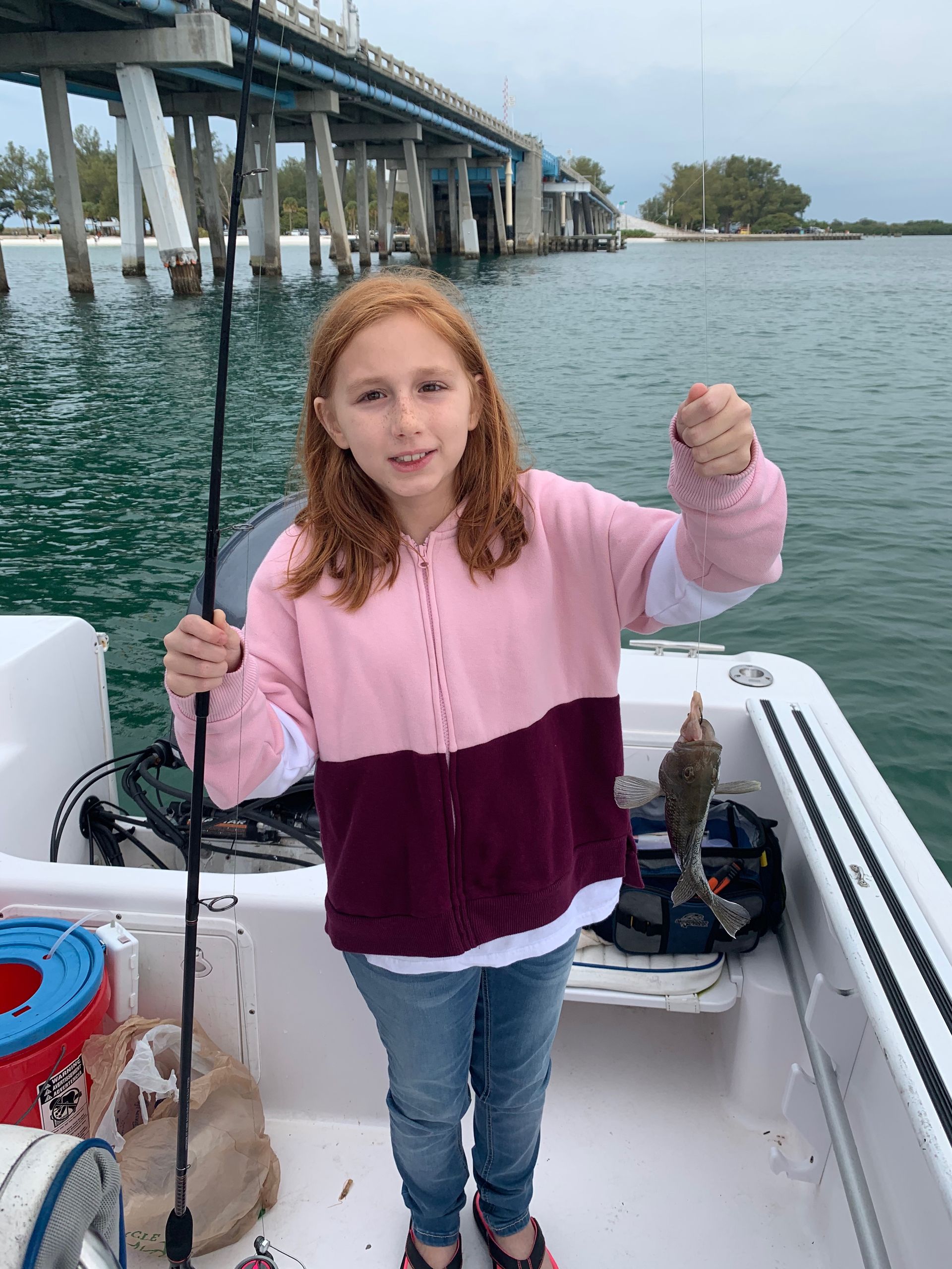 Girl on boat holds up small fish; pier in background.
