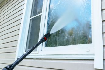 A person power washing a window on a house with white siding.