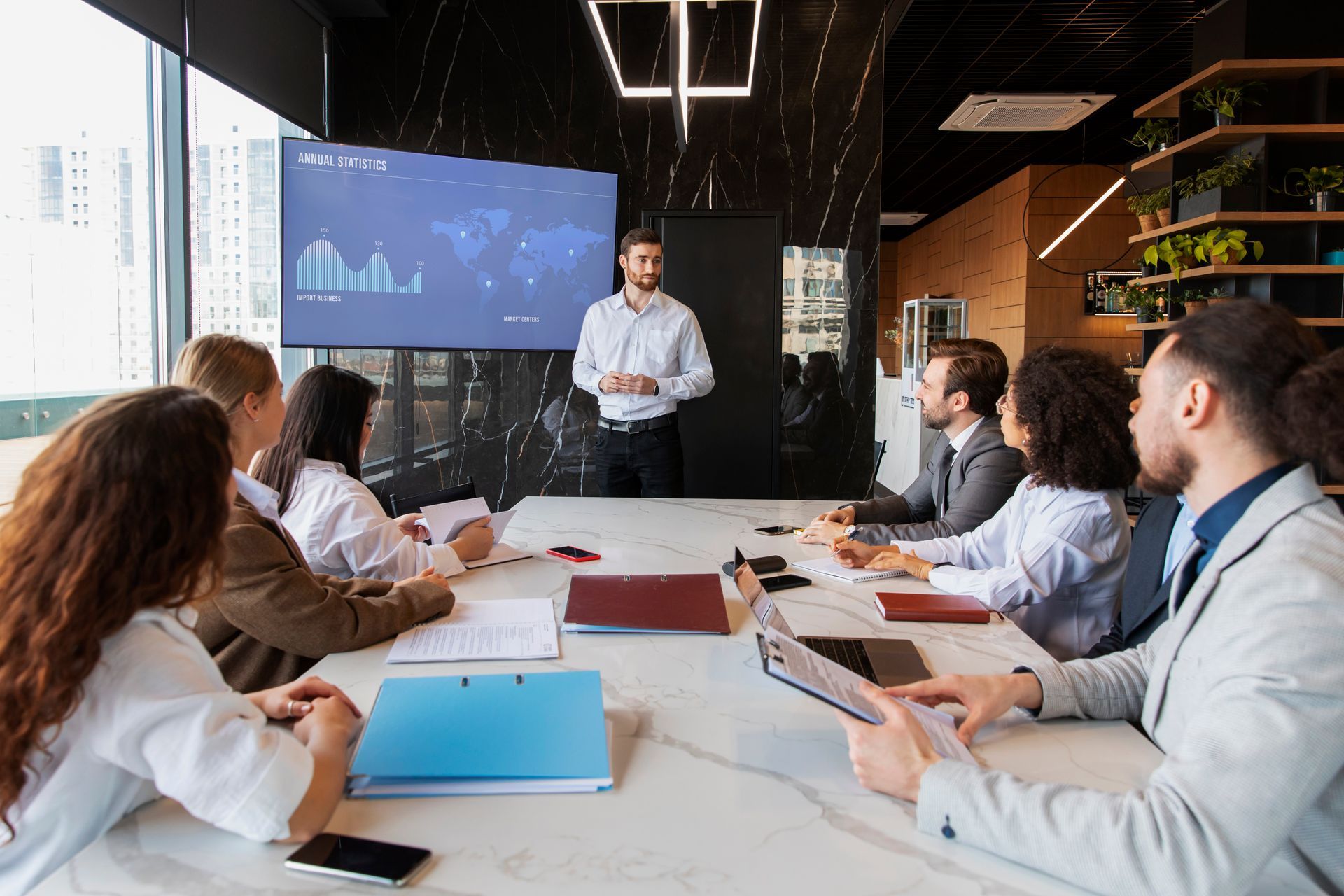 A man presenting at a conference table to a group of colleagues in a modern office; a screen displays a world map with data.