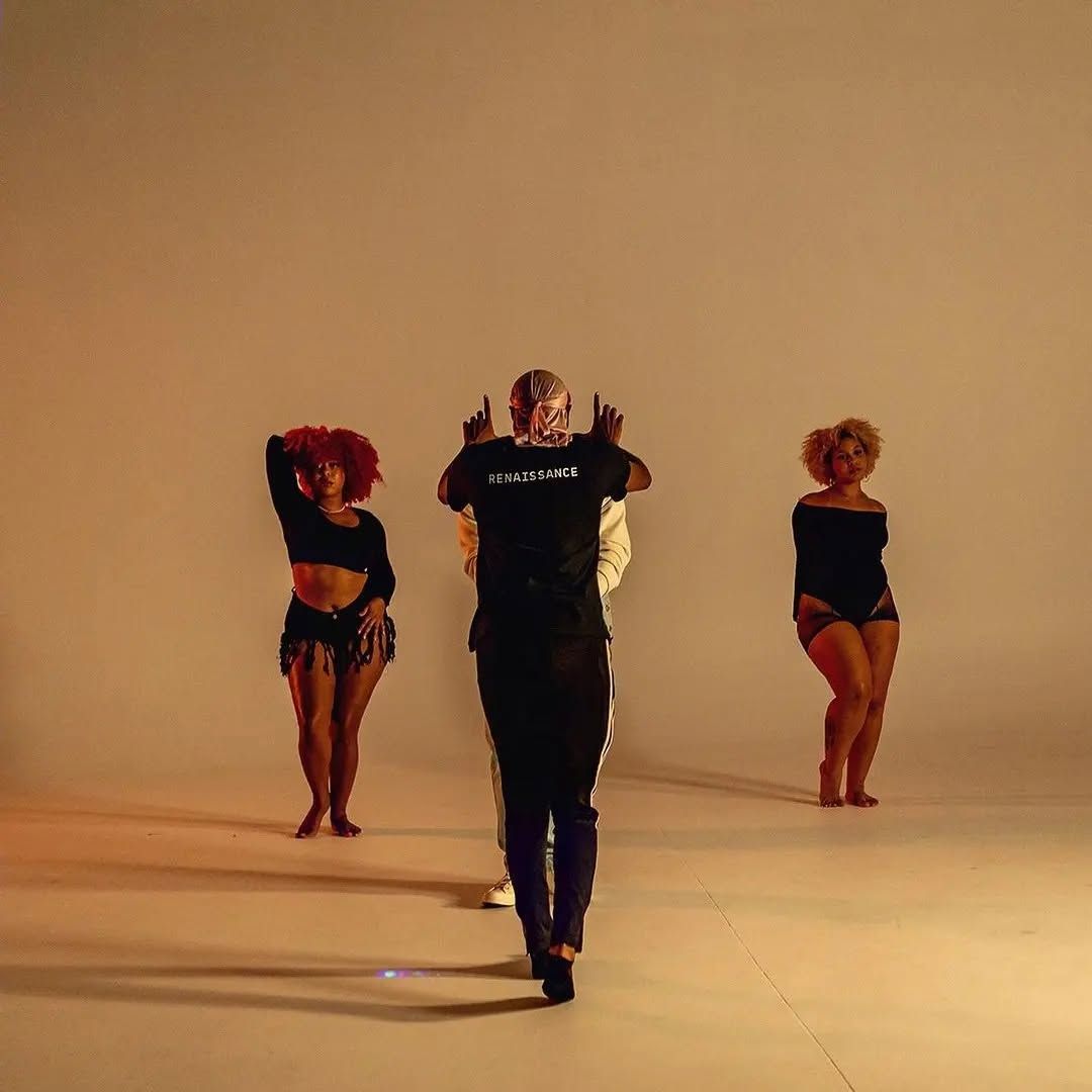 Three dancers in black outfits pose on a beige studio backdrop, center dancer facing forward.