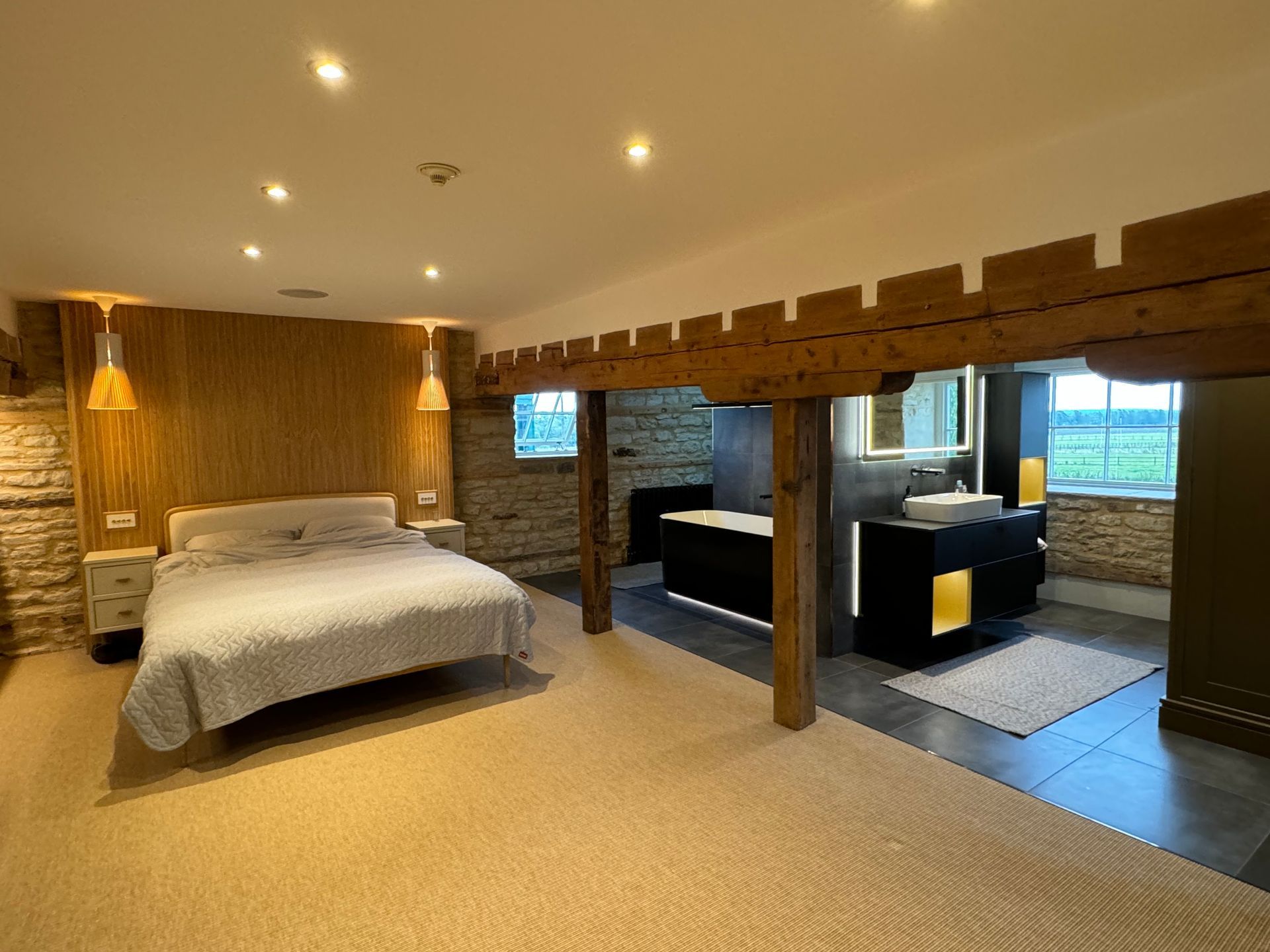 Bedroom with open bathroom. Features bed, exposed beams, stone walls, and modern vanity.