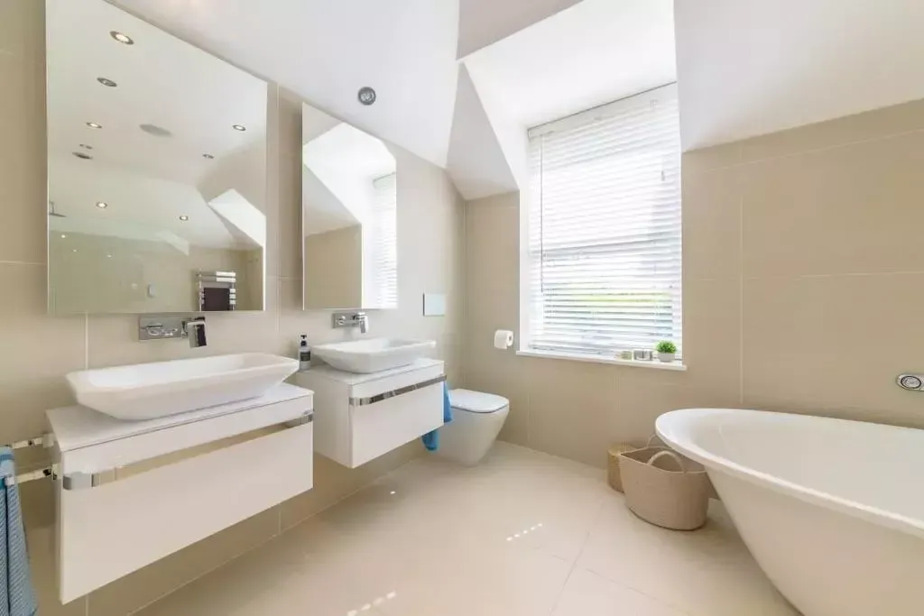 Bright bathroom with two sinks, a toilet, a bathtub, and a window with blinds.