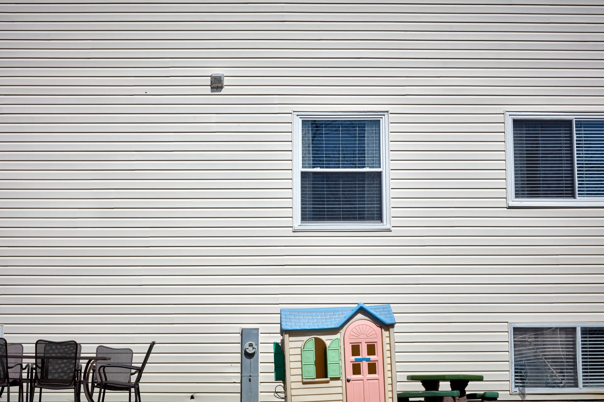 Doll House. The backyard of a modern family home awash with stark sunlight, amplified by the blinding reflection of vinyl siding. Streamwood, Illinois 2023 - Robert Swiderski