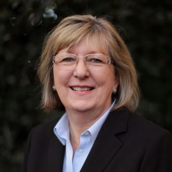 Smiling woman with glasses, light skin, short blonde hair, wearing a black blazer and blue shirt.