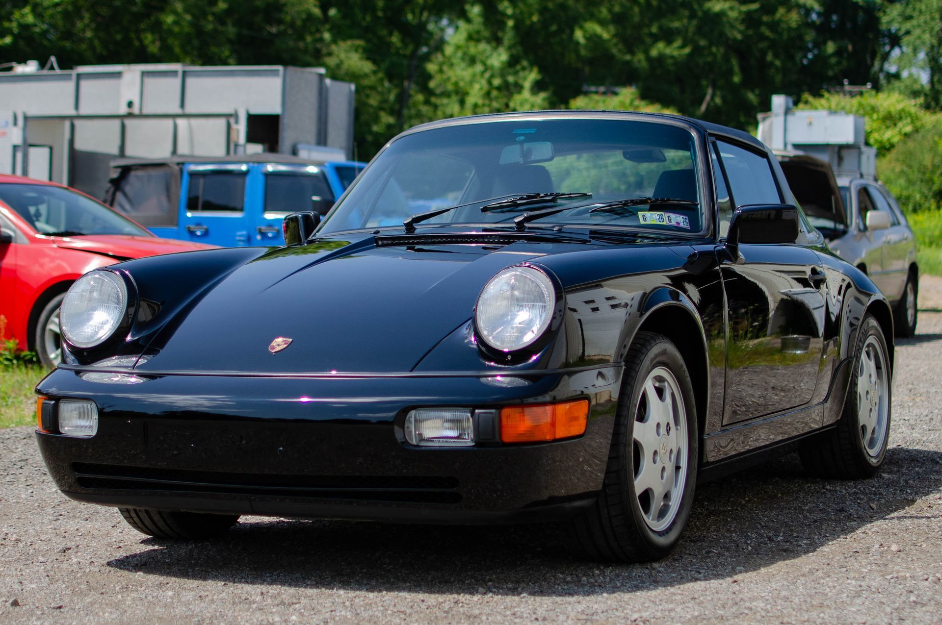 Black Porsche 911 parked on gravel, other cars in background.