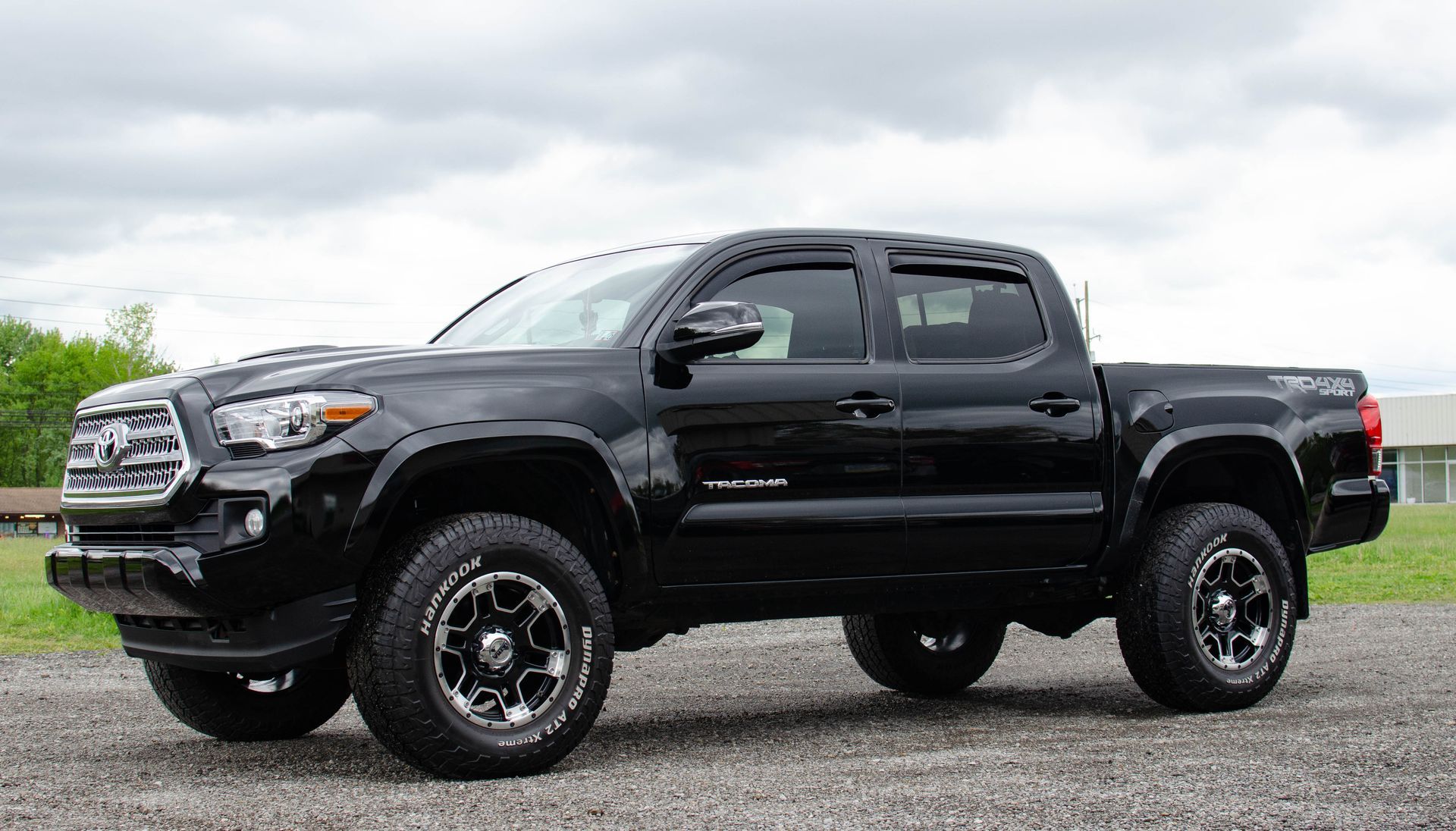 Black Toyota Tacoma pickup truck parked on gravel, cloudy sky in background.