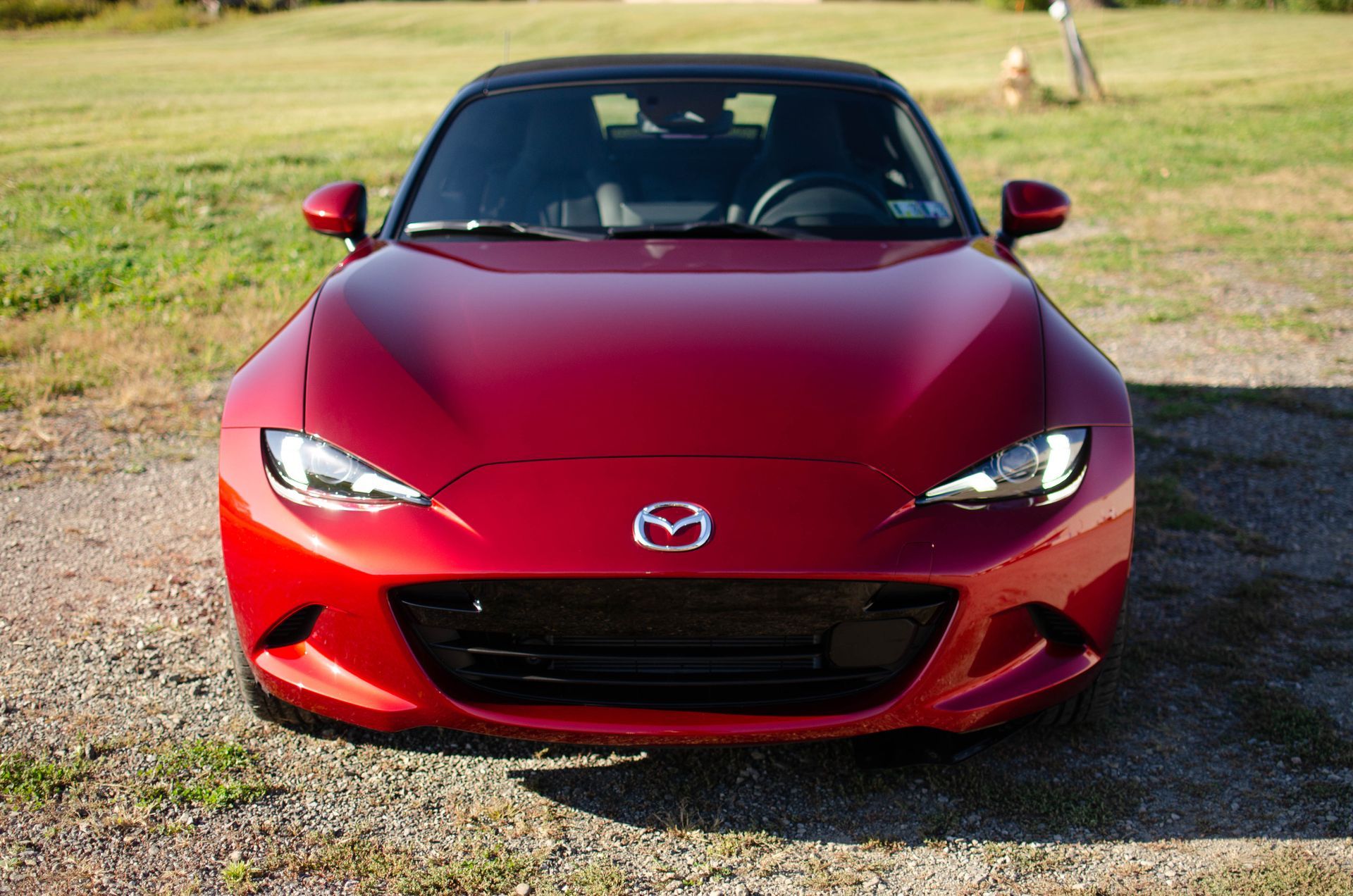 Red Mazda Miata convertible parked on gravel in front of a green field, top up.