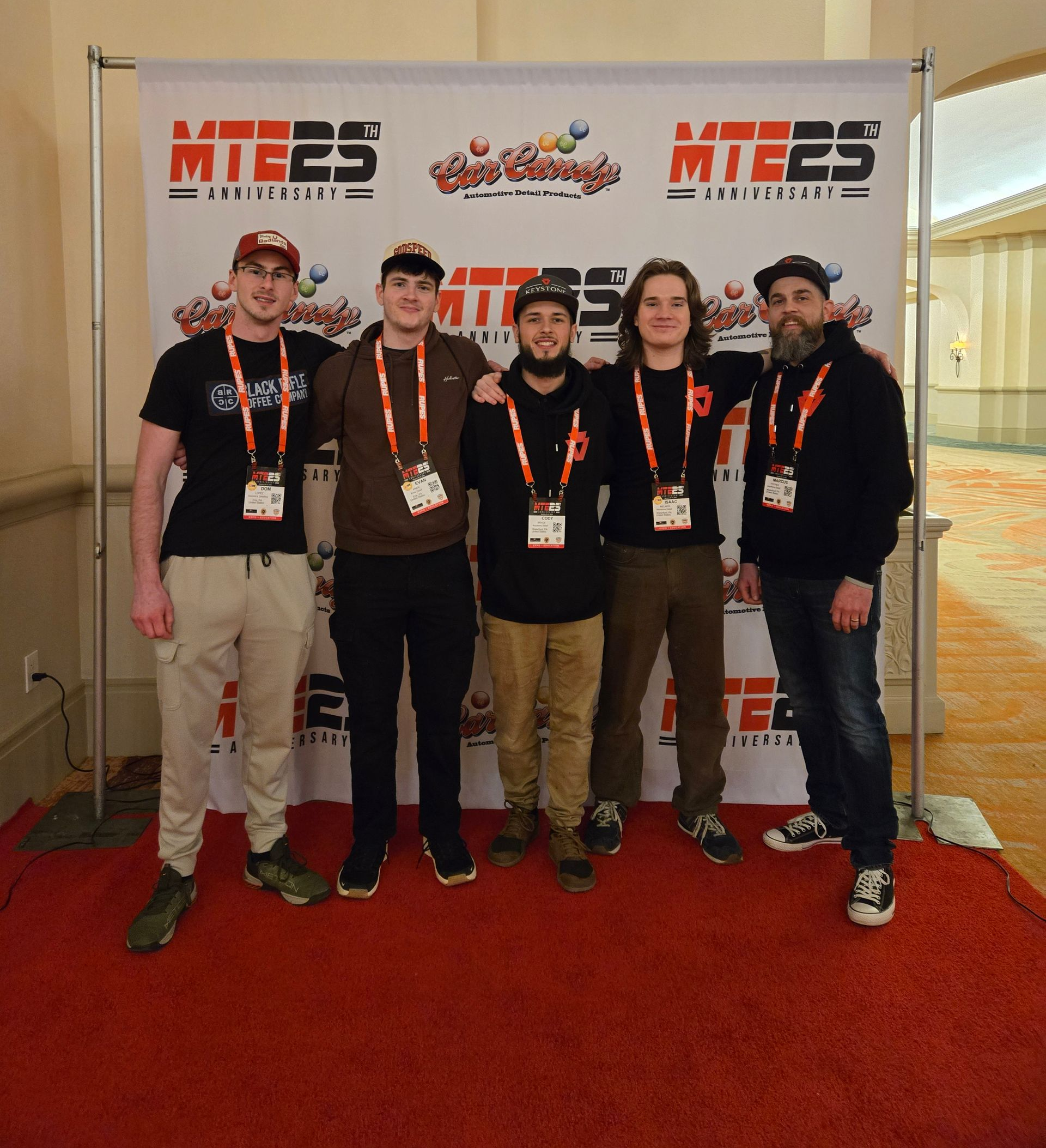 Man with beard, arms crossed, wearing a black hoodie and hat, in front of red graffiti.