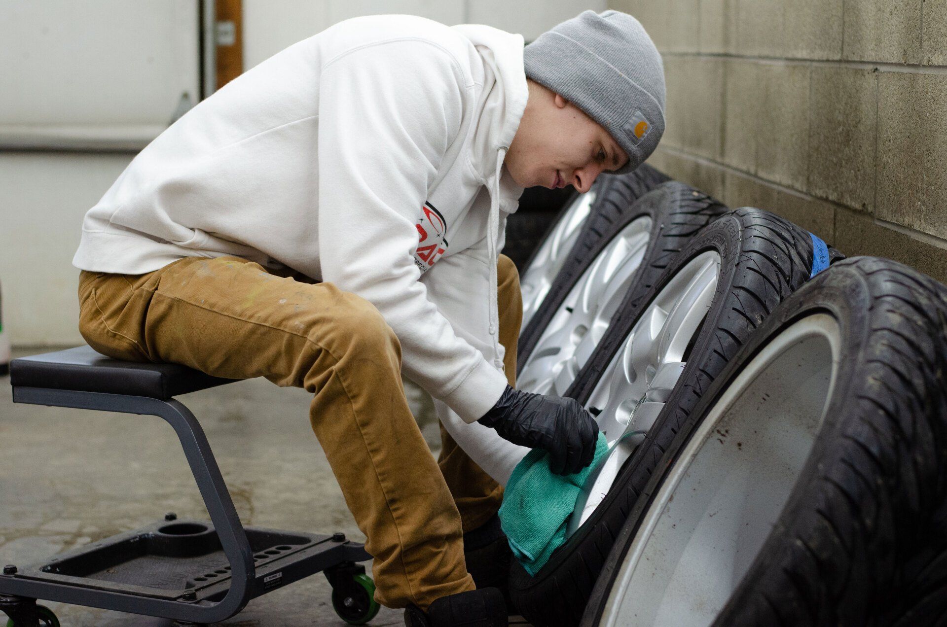 A man cleaning a car wheel in a garage, wearing a beanie, hoodie, and gloves.
