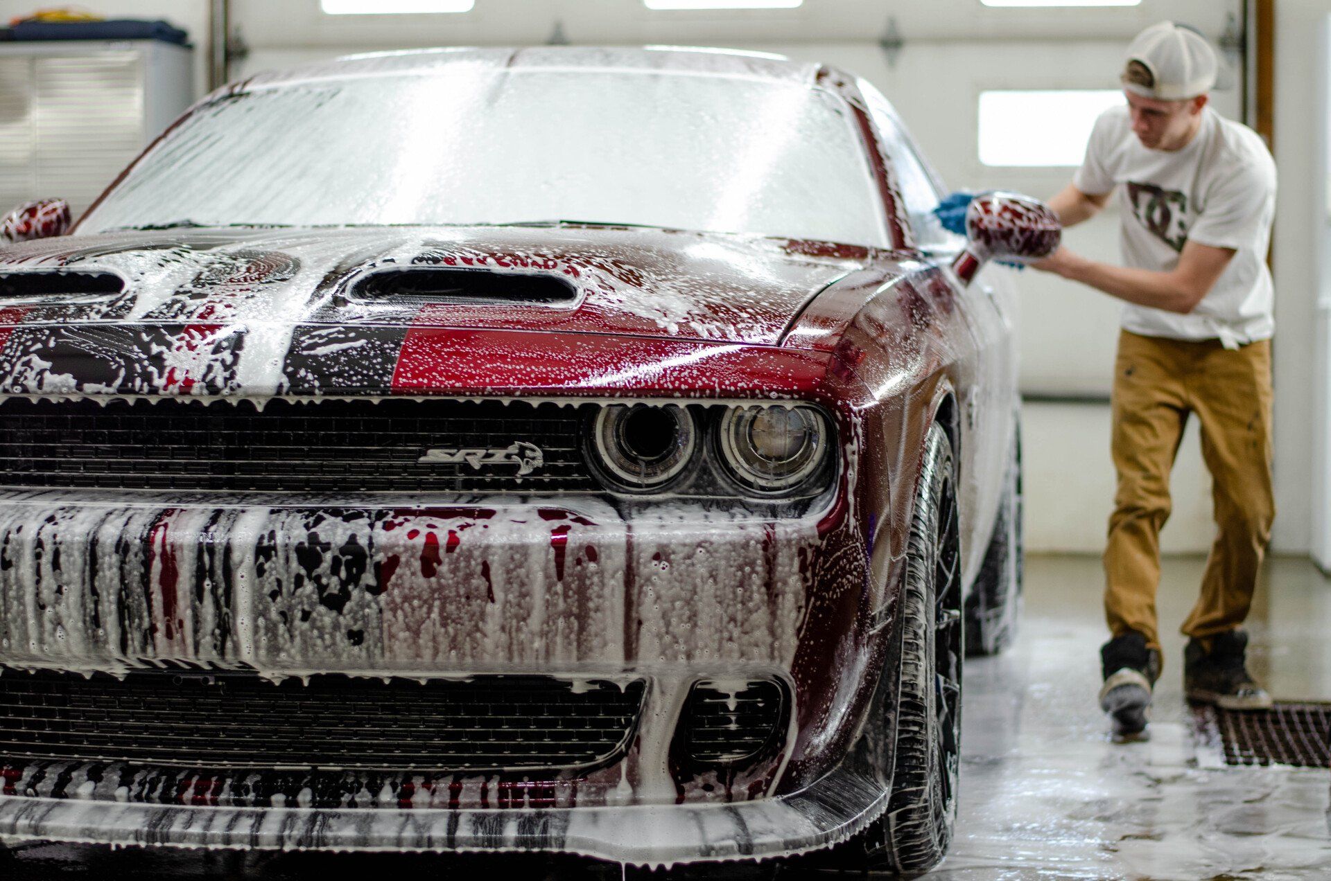 Man washing a red Dodge Challenger with soapy foam in a garage.