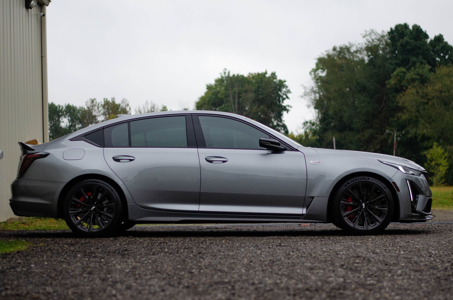 Silver Cadillac sedan parked near a building on a cloudy day. Black wheels and tinted windows.