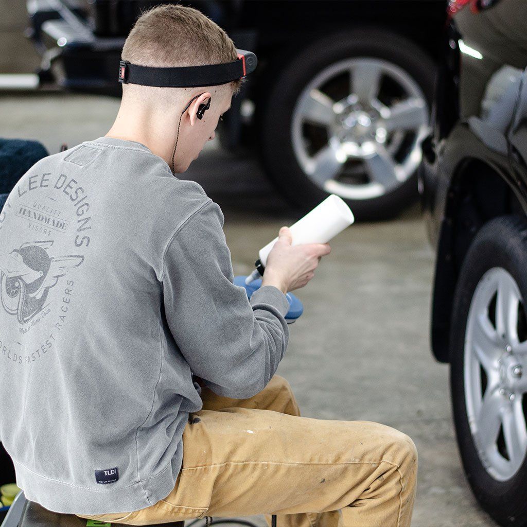 A man sits, working on a car. He wears a headlamp, gray sweater, and tan pants, holding a white object.