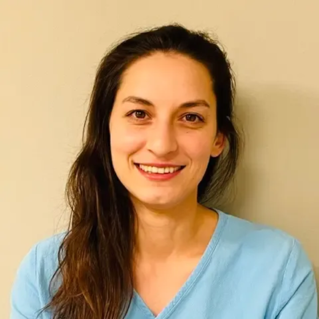 Kvinne med langt brunt hår smiler til kameraet, iført en lyseblå topp, mot en lys vegg.