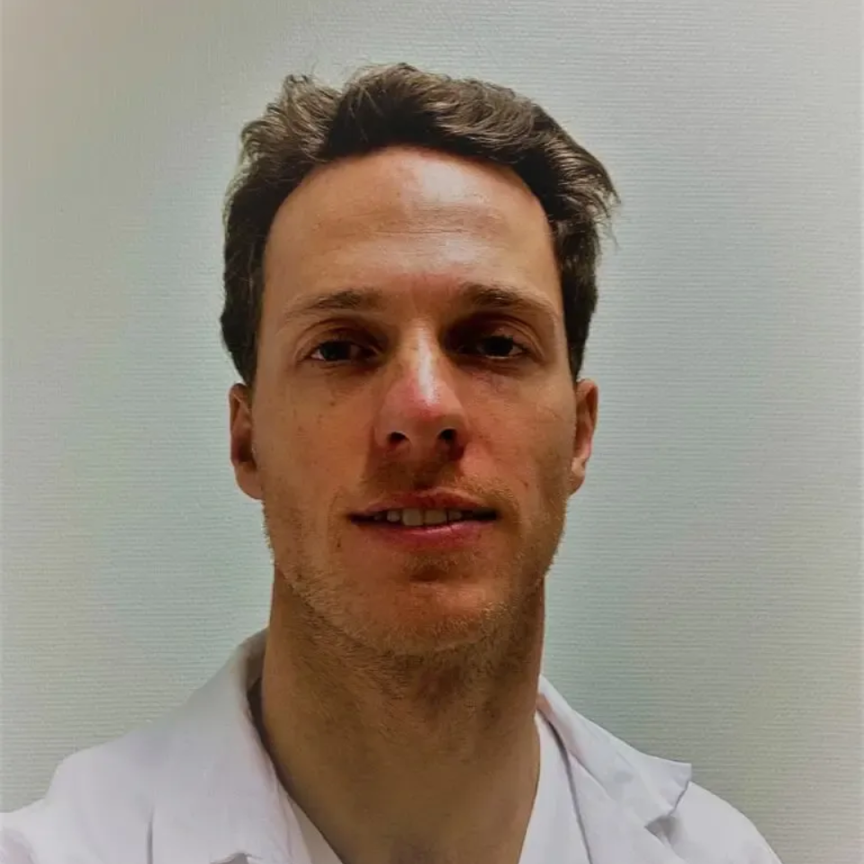 Mann i hvit frakk, smilende, mot en vanlig bakgrunn.