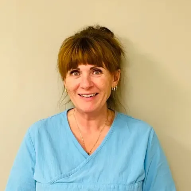 Kvinne med rødbrunt hår i en knute iført blå uniform, smiler til kameraet. Beige veggbakgrunn.