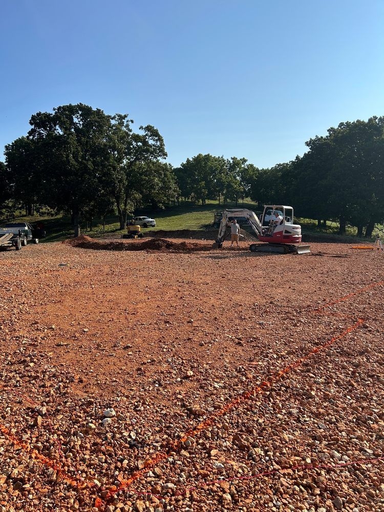 Construction site with excavators on a red dirt lot beside trees under a clear blue sky