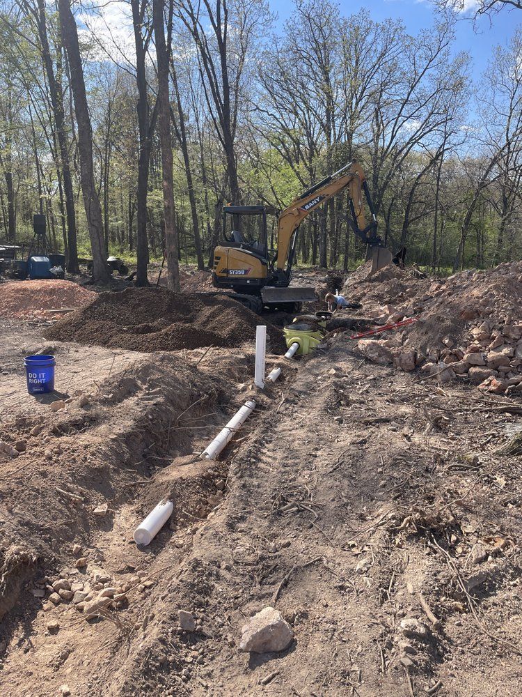 Excavator at a wooded construction site beside a dug trench with exposed pipes and dirt piles