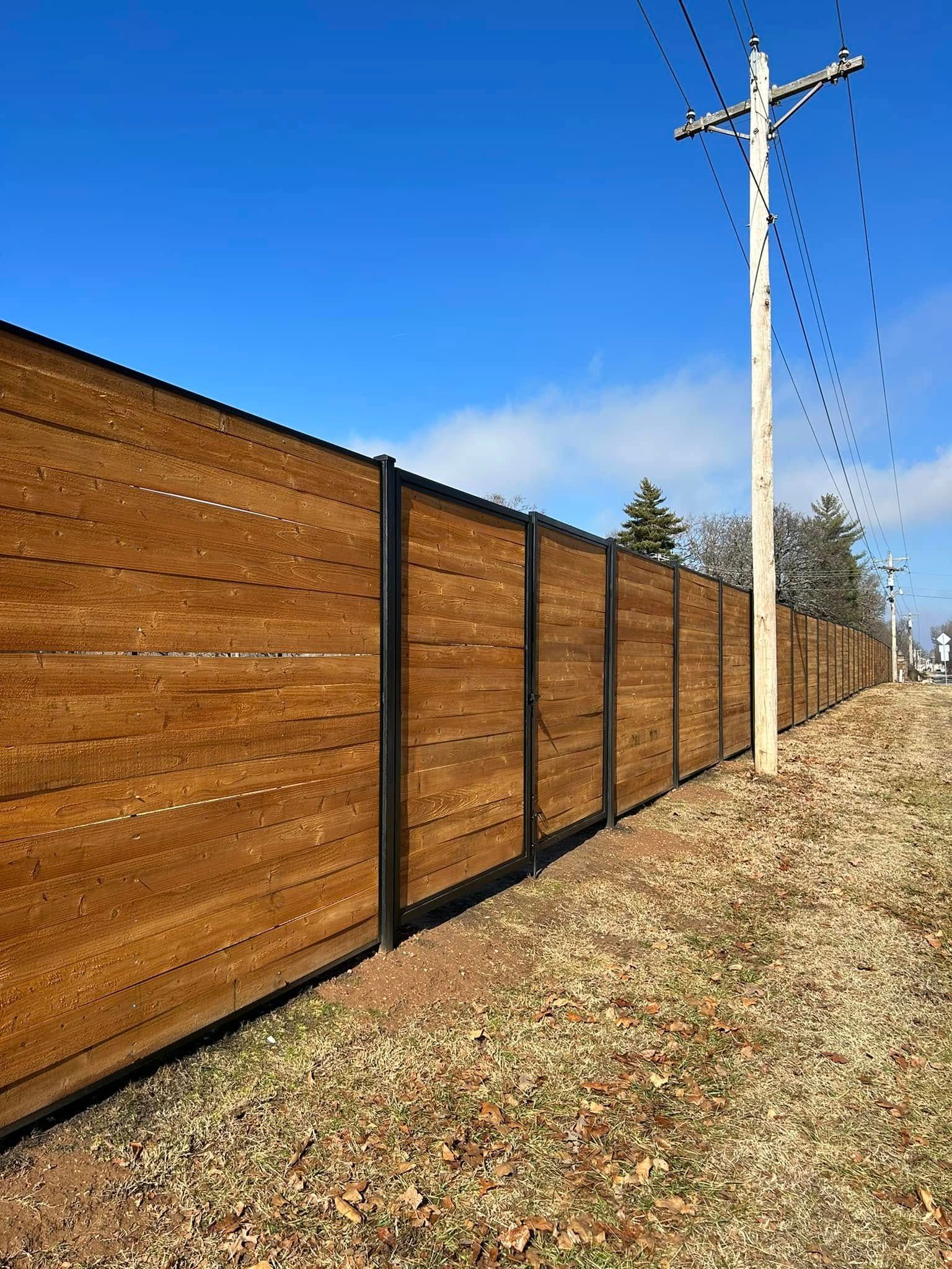 Wooden privacy fence along a gravel roadside under a clear blue sky with utility poles.
