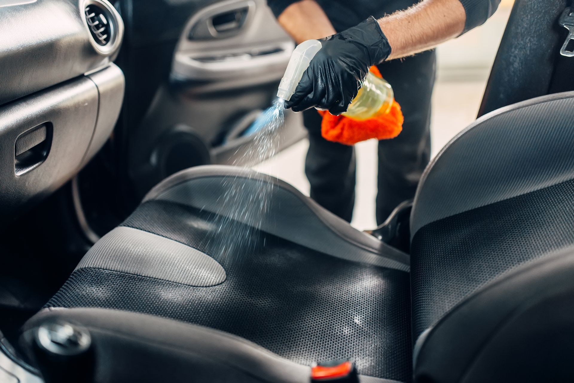 A person polishing a black Ford Mustang. They are wearing a black shirt, blue shorts, and a cap, working in a garage.