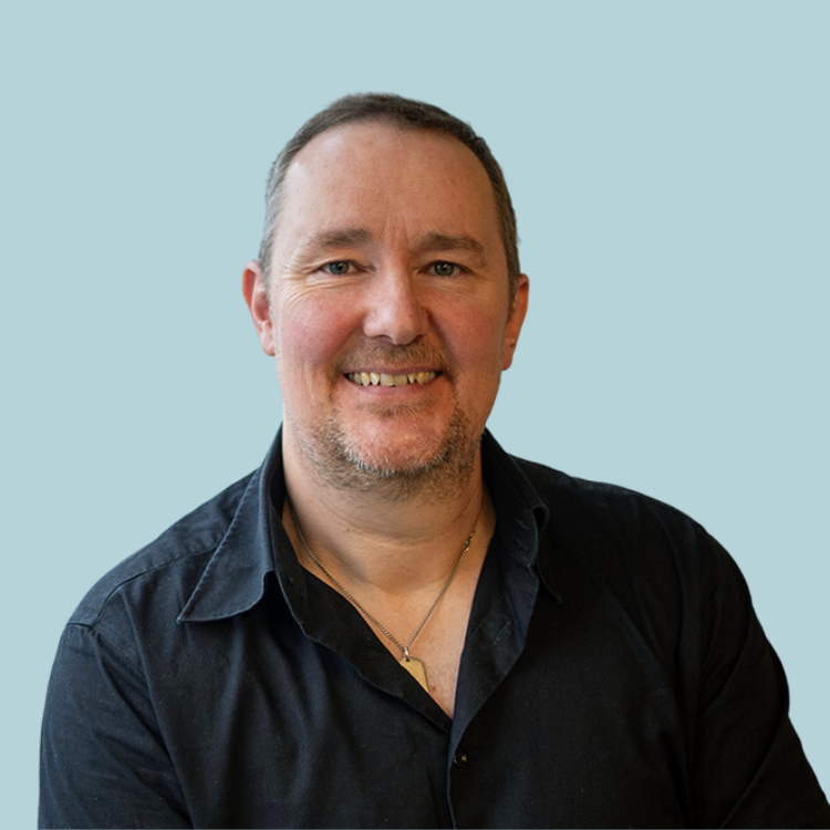 Man with short hair and stubble smiles, wearing a black shirt, against a light blue background.
